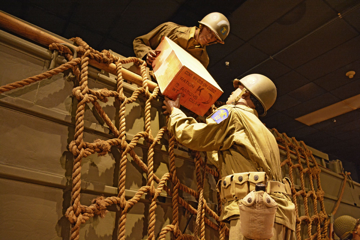 Two mannequin Soldiers handing off a wooden box to each other over a wall as part of a museum display at the US Army Quartermaster Museum.